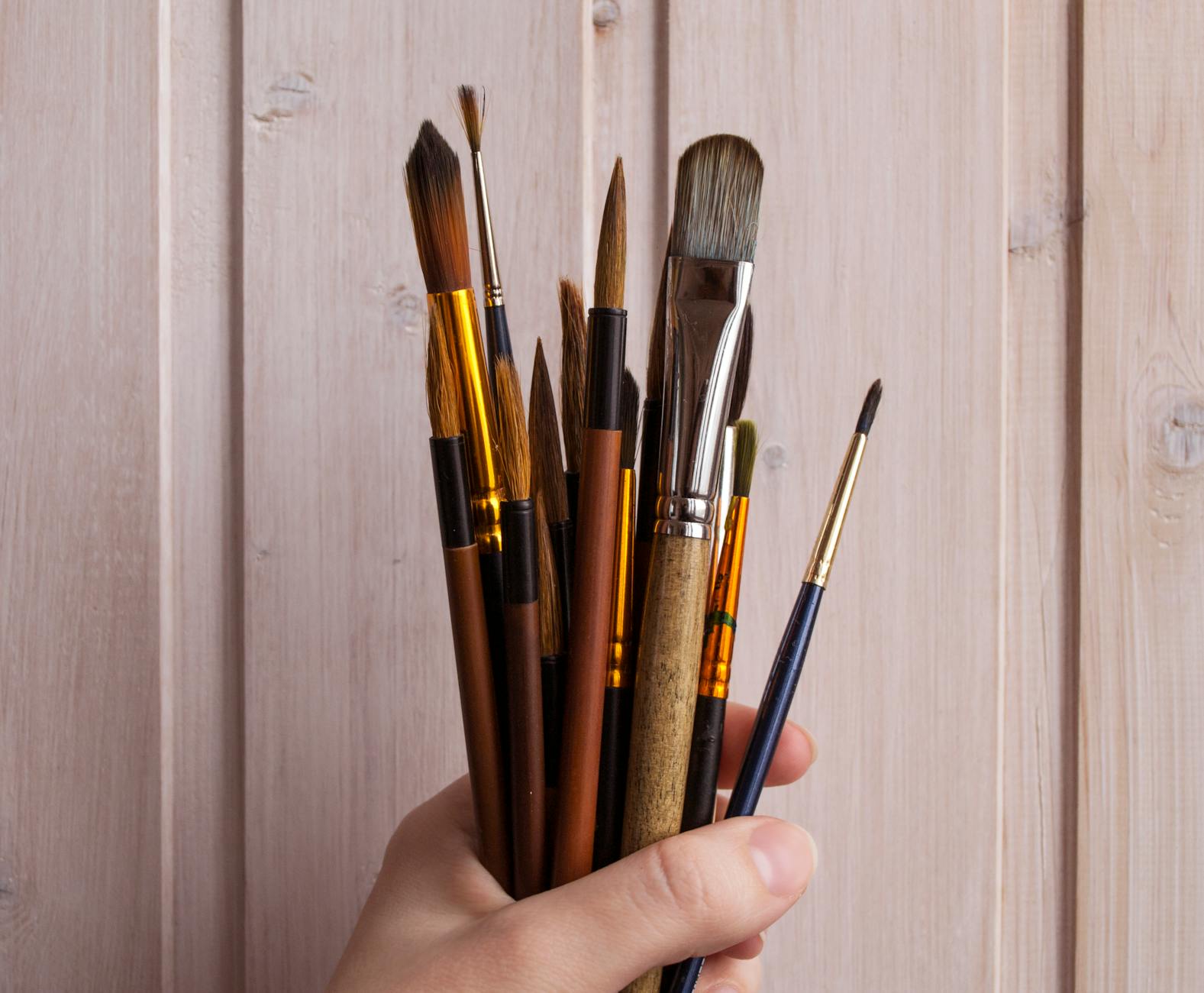 Efekt szczotkowanego drewna: jak go uzyskać domowymi narzędziami? Dłoń trzymająca pędzle malarskie na tle jasnej drewnianej deski