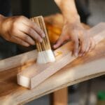 Czym zabezpieczyć drewno po szlifowaniu: olej, lakier, lazura czy wosk? Rzemieślnik nakłada olej pędzlem na oszlifowaną deskę drewnianą