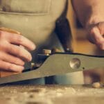 Dylatacje w konstrukcjach drewnianych: gdzie są potrzebne i jak je wykonać? Stolarz strugający drewnianą belkę w warsztacie