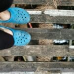 Jakie dylatacje zostawić przy podłodze drewnianej i dlaczego to ważne? Stopy w niebieskich sandałach na drewnianym pomoście nad wodą